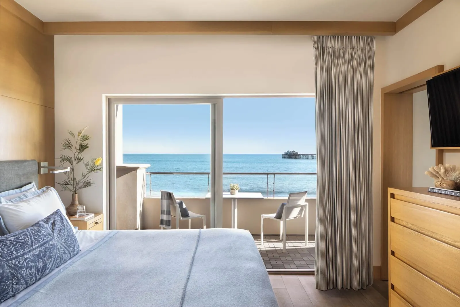 One Bedroom Ocean Front Suite at Malibu Beach Inn: a bed beside a sliding glass door onto a balcony with a small table and two chairs, and the sea beyond