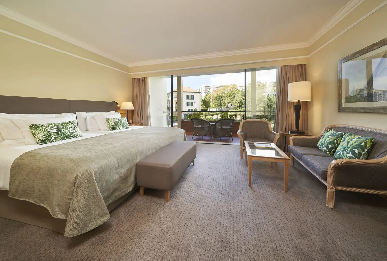 Standard at The Cliff Bay, by PortoBay: a wide twin-bed pair made up as one bed, a sofa and armchair, and a balcony with two wicker chairs looking onto trees and a neighbouring building