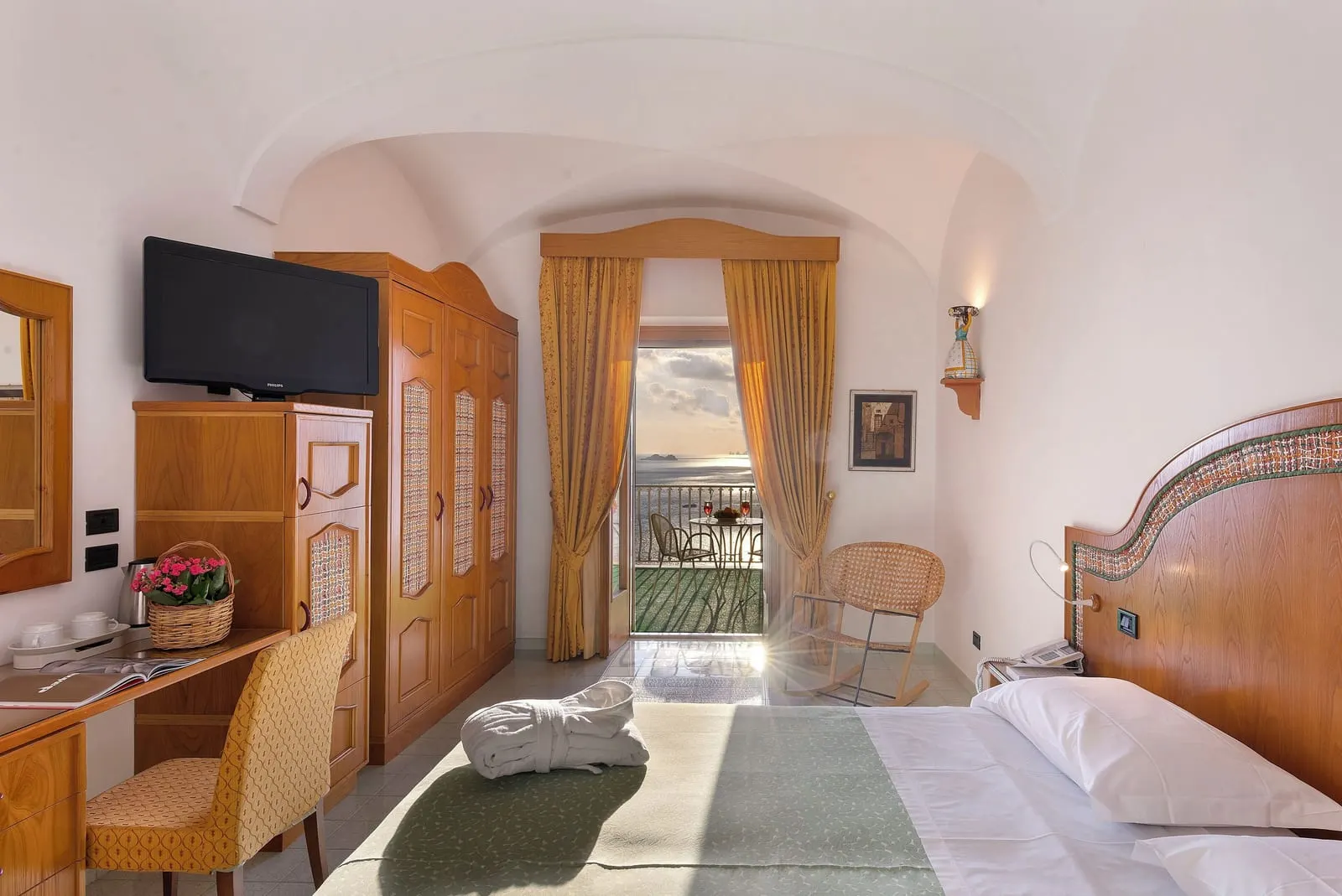 Classic Room with sea view terrace all'Hotel Tramonto d'Oro: la vista dal letto, oltre una sedia a dondolo, fino alle porte aperte sulla terrazza, con un tavolo e sedie all'esterno e il mare in lontananza.