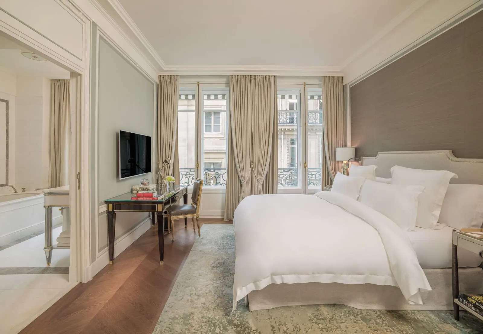 Premier Room at Hôtel de Crillon, A Rosewood Hotel: a king bed against a taupe fabric wall, a writing desk under a wall-mounted television, tall French windows with iron railings, and the bath through a doorway on the left