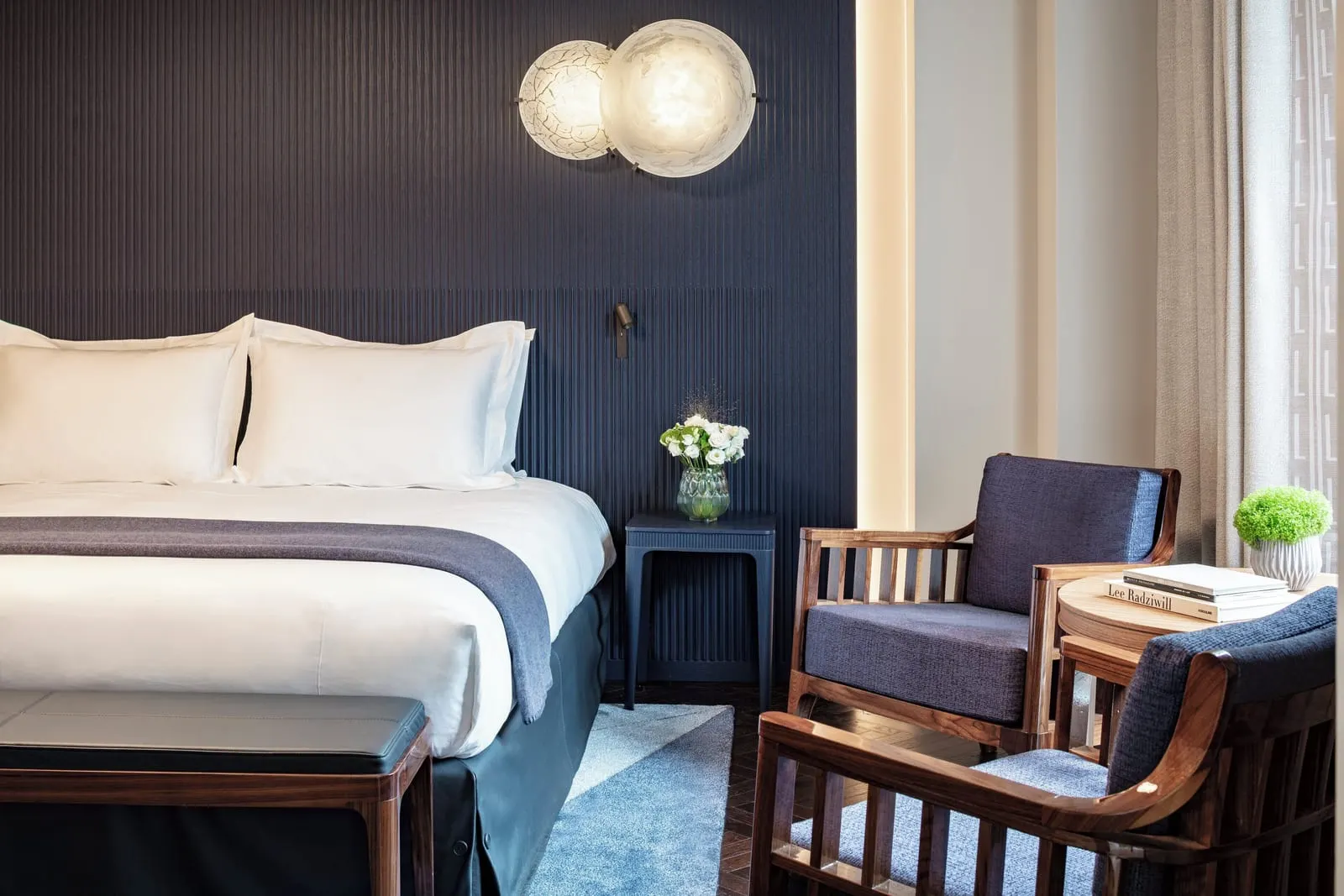 Family Room at Mandarin Oriental Lutetia, Paris: a double bed against a dark blue fluted headboard with two round glass wall lights, a bench at its foot and two blue armchairs by a side table