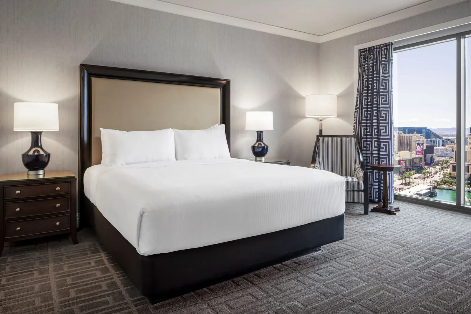 Palace Partial Strip View Room im Caesars Palace: ein Kingsize-Bett mit dunklen Lampen, ein gestreifter Sessel und ein Beistelltisch an einem hohen Fenster