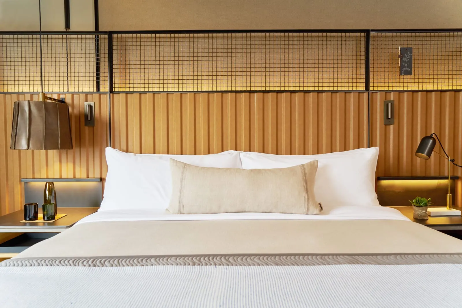 Dumbo King im 1 Hotel Brooklyn Bridge: ein Kingsize-Bett mit weißen Kissen und einem langen Leinenkissen unter einem gerippten hellbraunen Kopfteil, mit einer Lampe mit Lederschirm und einer schwarzen Schreibtischlampe auf den Nachttischen