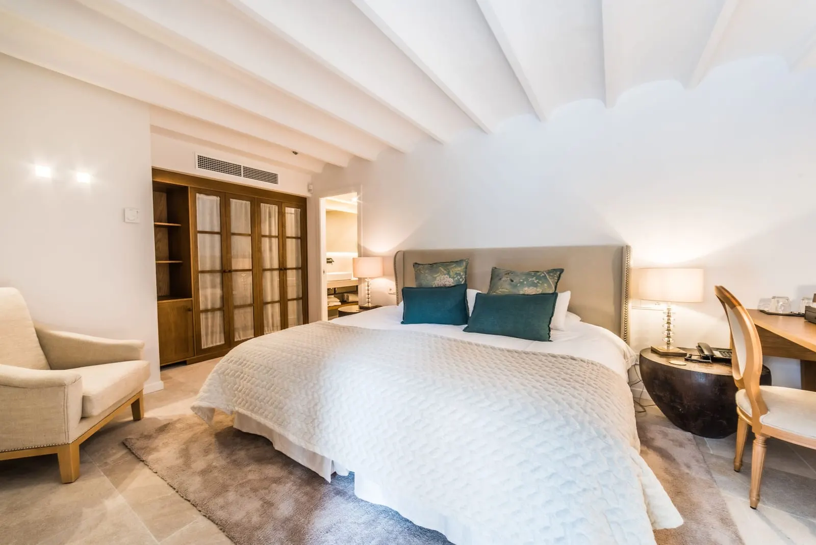 Double Room Garden at L'Avenida Hotel: a double bed with an upholstered headboard under a white beamed ceiling, with an armchair, a desk and a glazed wardrobe
