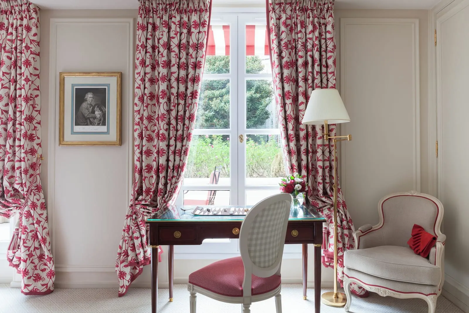 Deluxe Room with balcony at Le Bristol Paris: a writing desk set with a backgammon board, an armchair and a brass floor lamp in front of French windows hung with red floral curtains