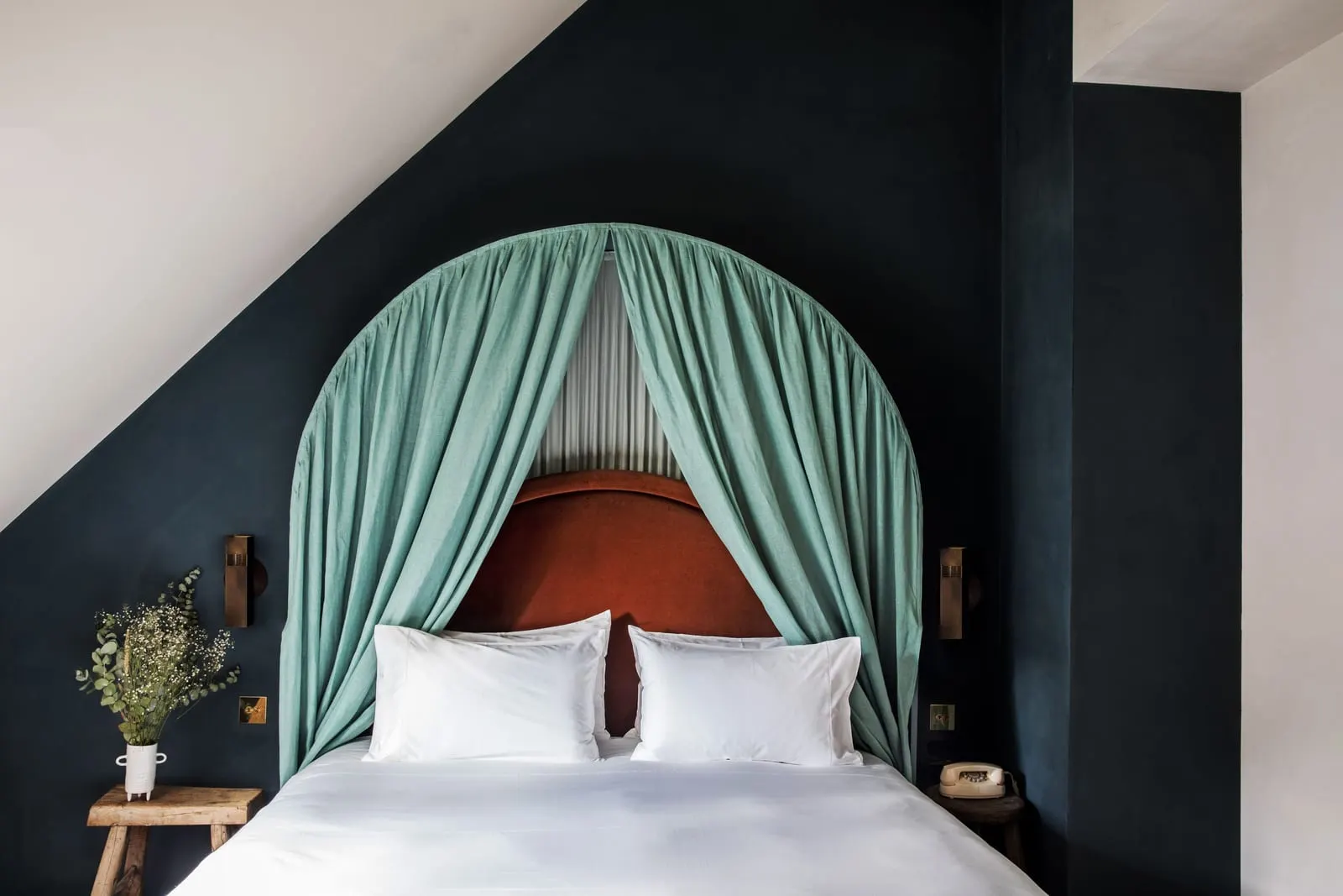 Parisian Eaves at Grands Boulevards Experimental: a bed framed by an arched green canopy and a rust velvet headboard against a dark blue wall that follows the slope of the roof, with wooden stools as bedside tables