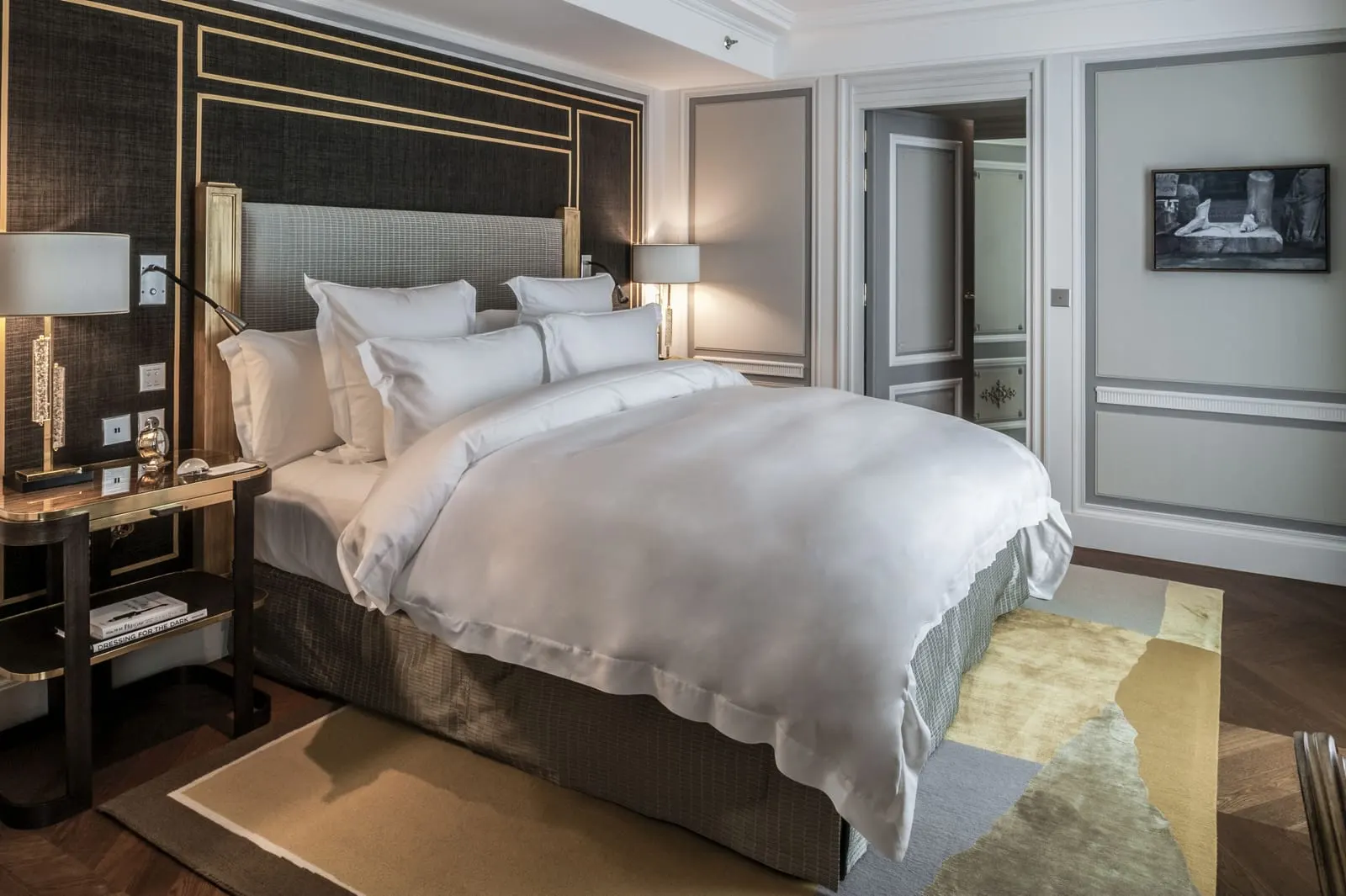 Grand Premier Room at Hôtel de Crillon, A Rosewood Hotel: a king bed with a checked headboard set into a dark wall lined in brass, a brass and wood bedside table, and grey panelled walls with a door standing open