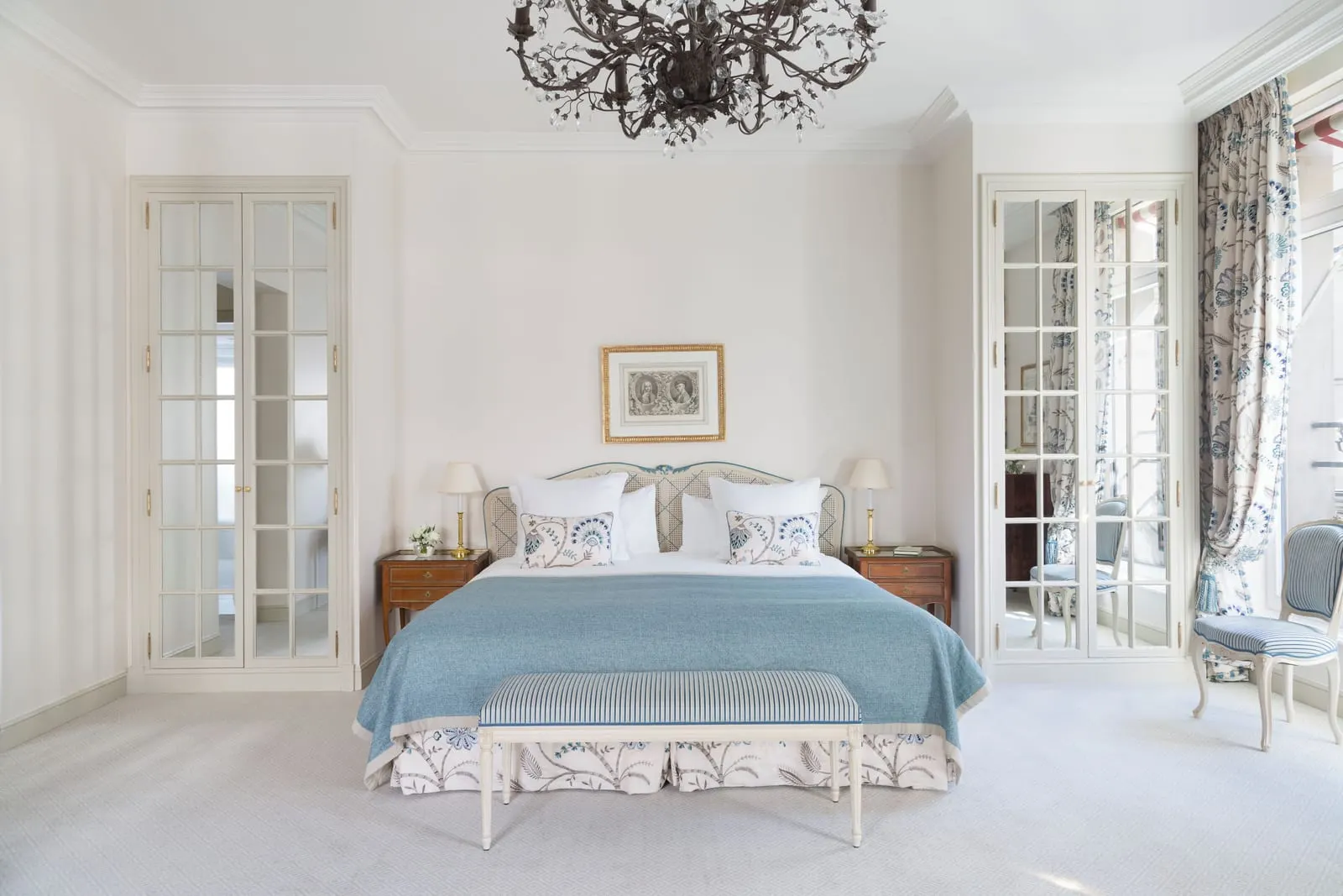 Corner Junior Suite at Le Bristol Paris: a bed with a pale blue throw and a striped bench between two pairs of mirrored doors, under a dark iron chandelier
