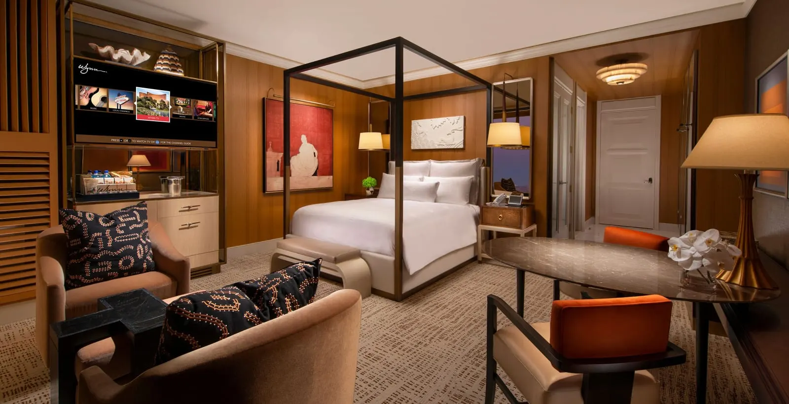 Wynn Tower Suite King at Wynn Las Vegas: a king bed under a dark canopy frame with a bench at its foot, two armchairs, an oval table with orange-backed chairs and a cabinet holding the television and minibar