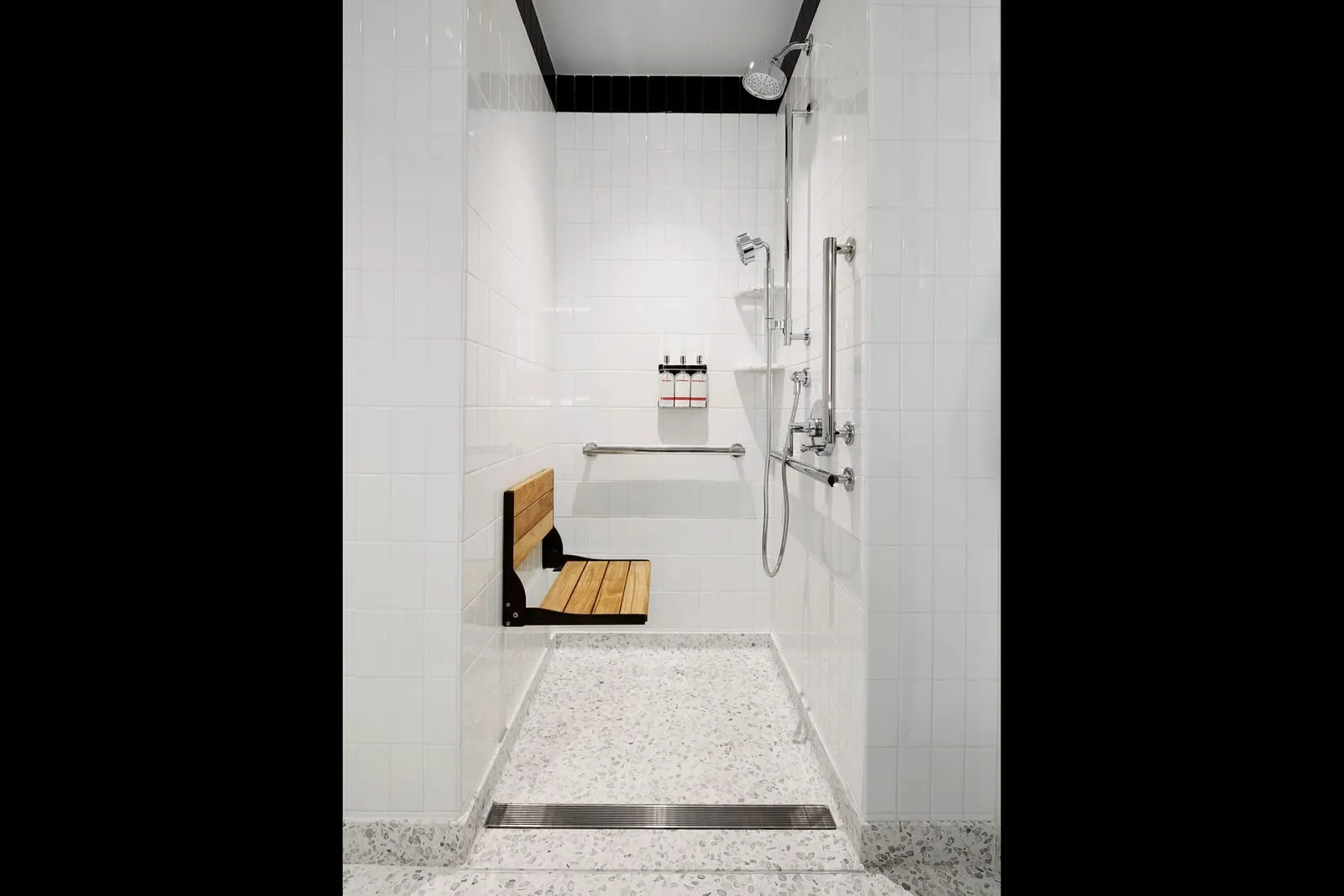 Accessible Deluxe King at TWA Hotel: a shower with a fold-down wooden seat, grab bars and a handheld shower on a rail, set in white tile with a terrazzo floor