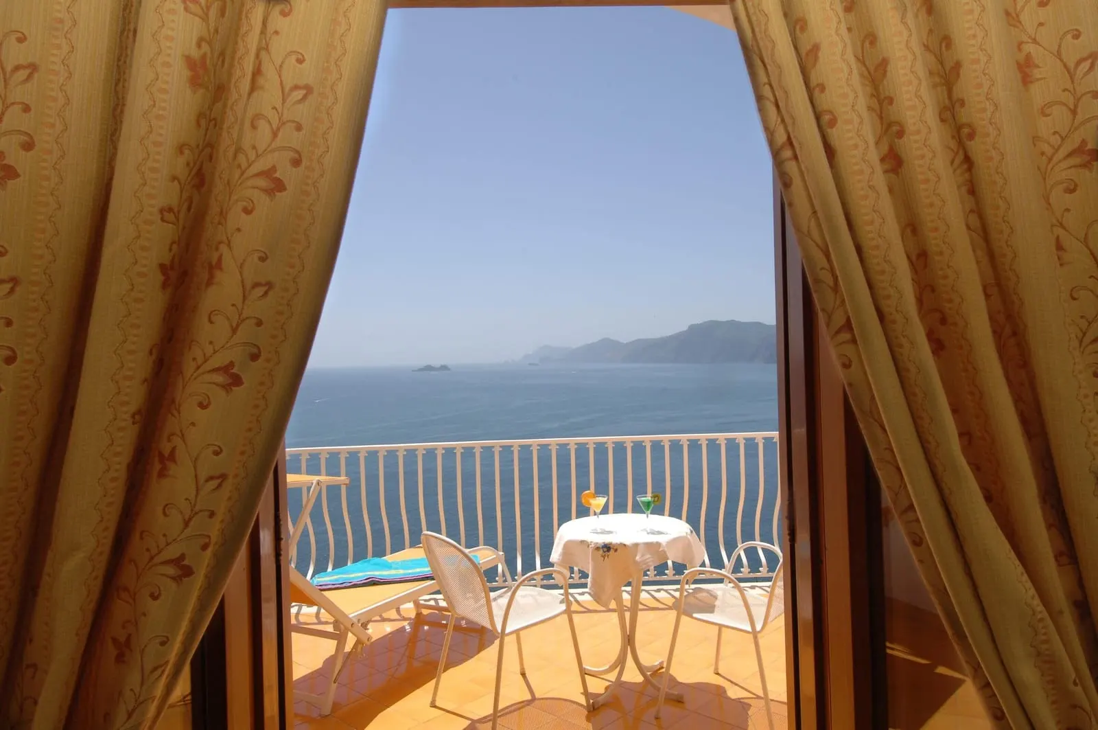 Classic Room with sea view terrace all'Hotel Tramonto d'Oro: porte con tende aperte su una terrazza piastrellata con un lettino, un tavolo e due sedie sopra il mare.
