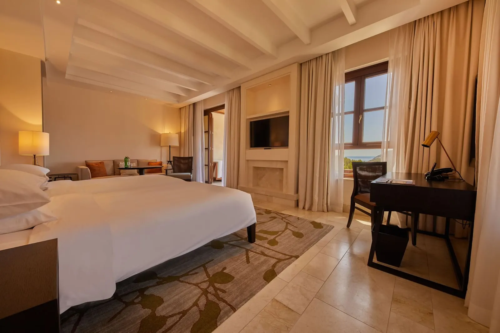 Junior Suite Resort View at Cap Vermell Grand Hotel: a large white bed under pale beams, with a sofa, a dark wood desk and a window