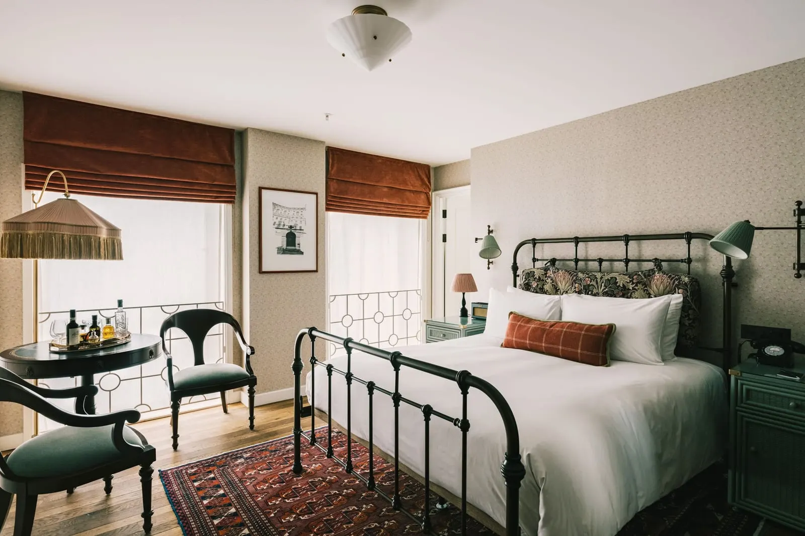 Cosy room at The Ned: an iron bed, a small round table with two chairs and windows with Roman blinds