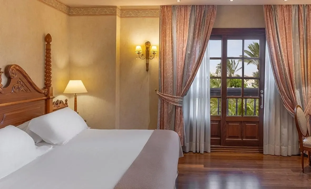 Deluxe double room with view of golf course at Elba Palace Golf Boutique Hotel: a carved wooden king bed beside glazed doors that open onto a balcony with palm trees beyond