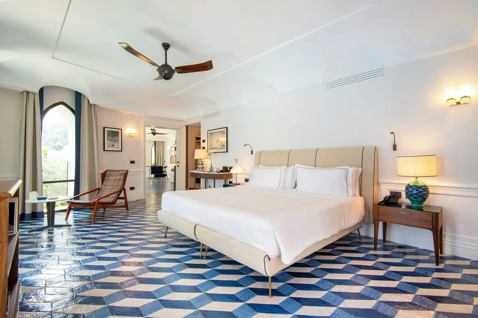 Corner Suite Sea View at Borgo Santandrea: a king bed under a ceiling fan, with an armchair by an arched window and the sitting room through a doorway.