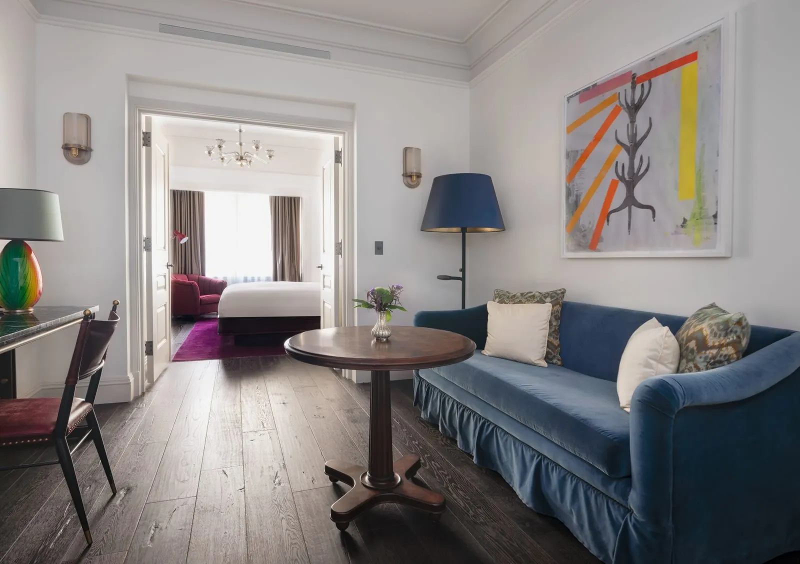 One Bedroom Suite at The Beekman: a sitting room with a blue velvet sofa, a round table and large abstract art, and the bedroom visible through a wide doorway