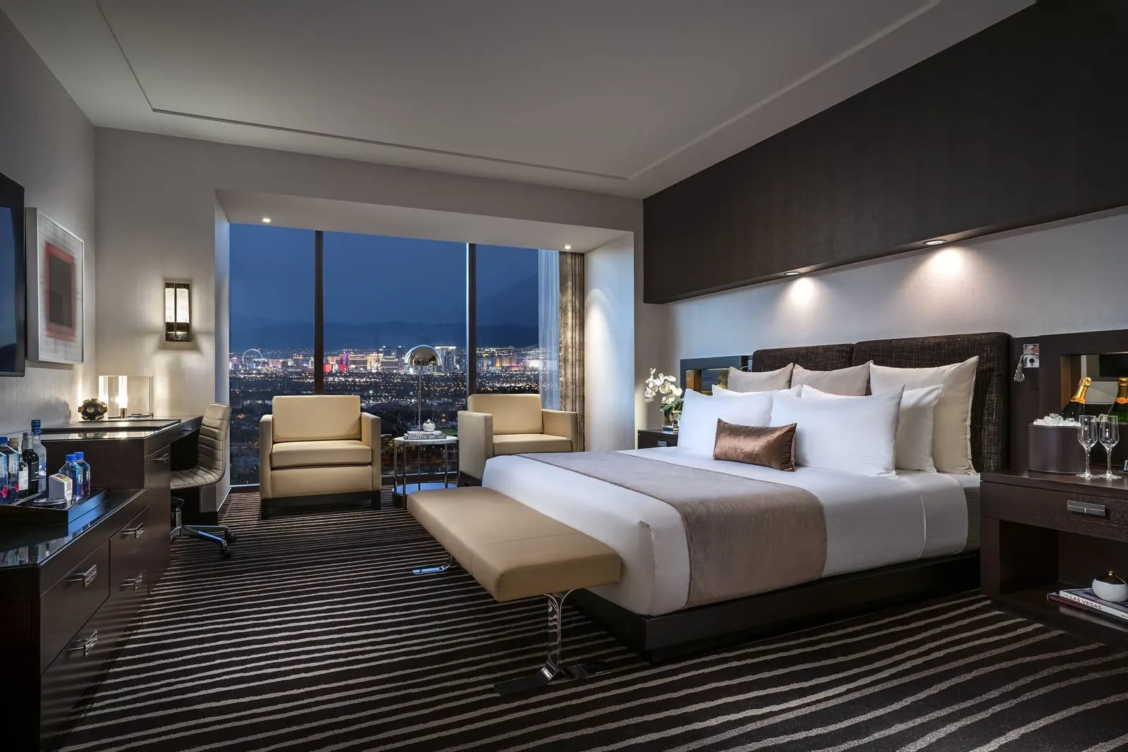 Strip View King at Red Rock Casino Resort & Spa: a king bed with an upholstered bench at its foot, two armchairs by a floor-to-ceiling window, and a desk and dresser along the left-hand wall
