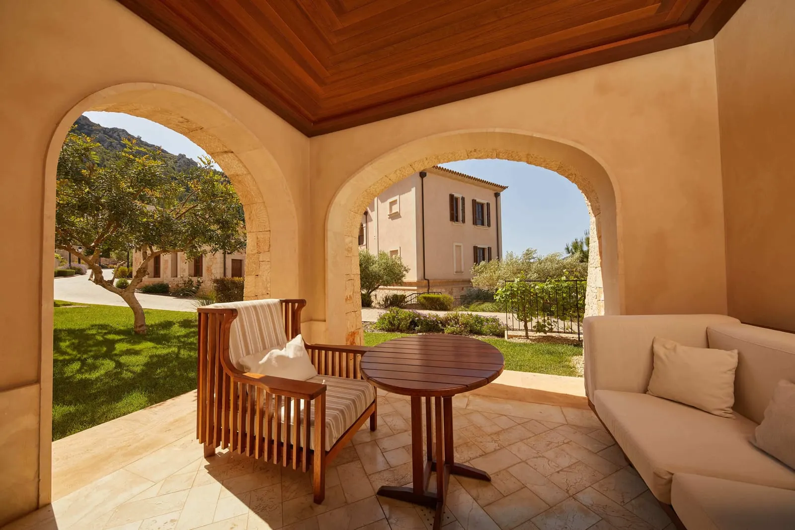 Junior Suite Garden at Cap Vermell Grand Hotel: a covered terrace with a timber armchair, a round table and a cushioned sofa, opening onto a lawn and a tree