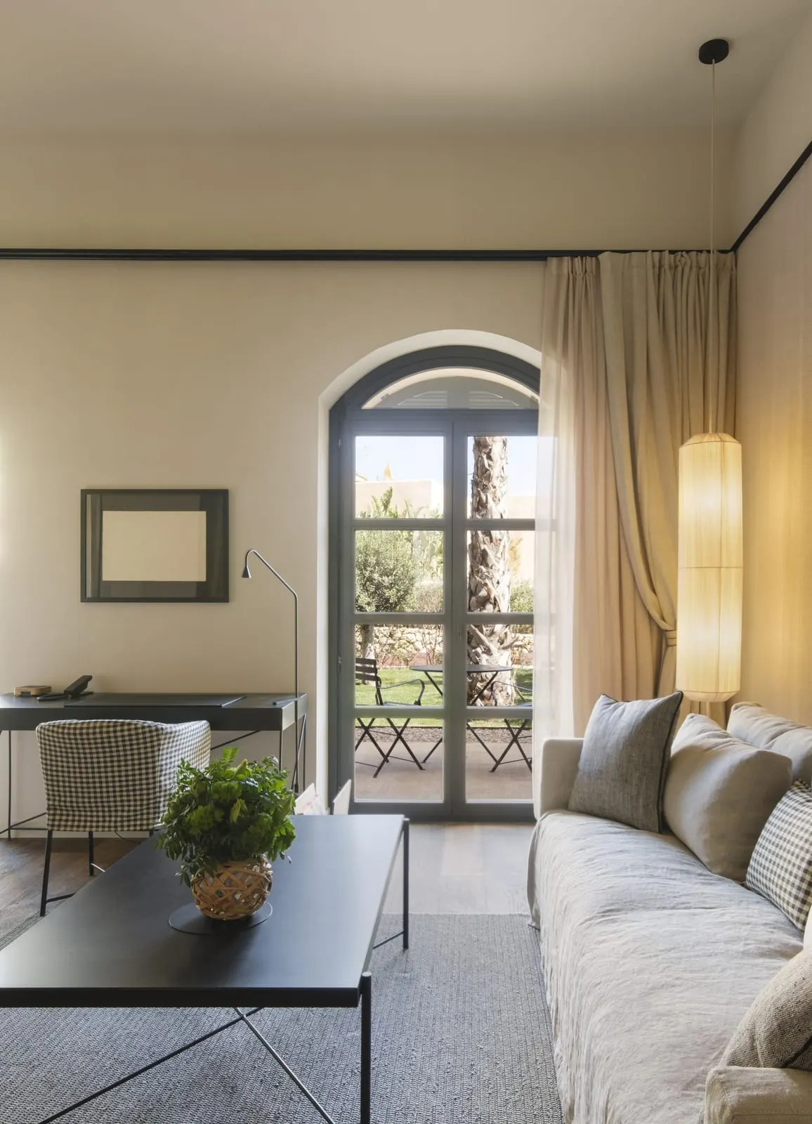 Terrace Suite at Can Ferrereta: a sitting area with a linen sofa and a desk, and a tall arched glass door opening onto a garden terrace with a table and chairs