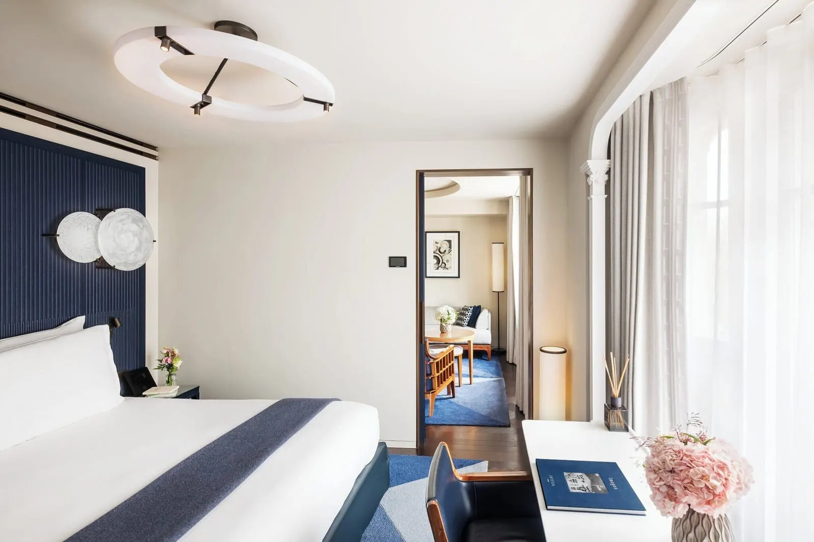 Deluxe Suite at Mandarin Oriental Lutetia, Paris: a double bed against a dark blue fluted headboard and a writing desk by sheer curtains, with a door open to the living room beyond