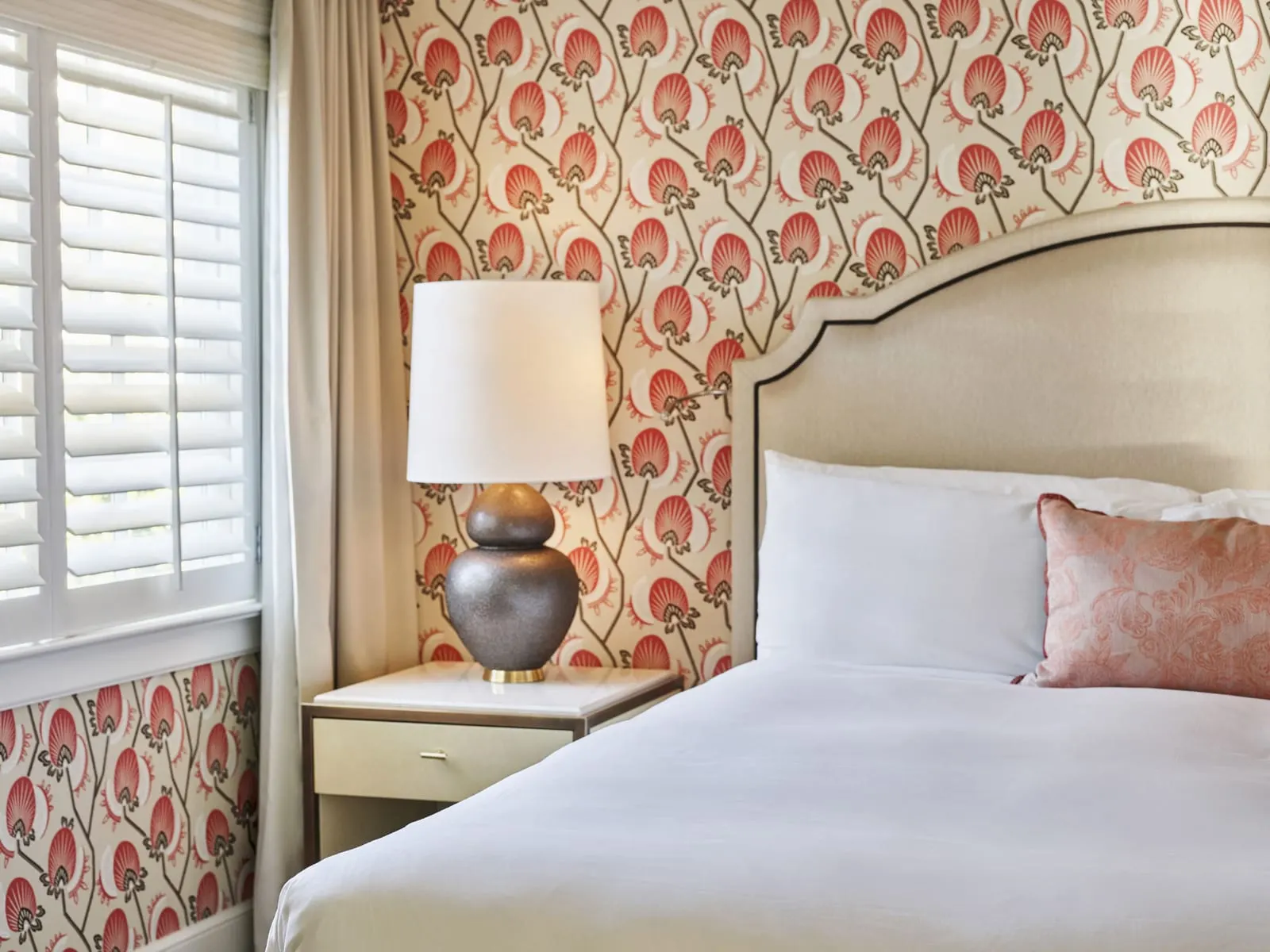 Bungalow Room at The Beverly Hills Hotel: the head of a bed with a pale upholstered headboard and a coral pillow, a speckled grey lamp on a bedside table, a coral and cream patterned wall and white window shutters