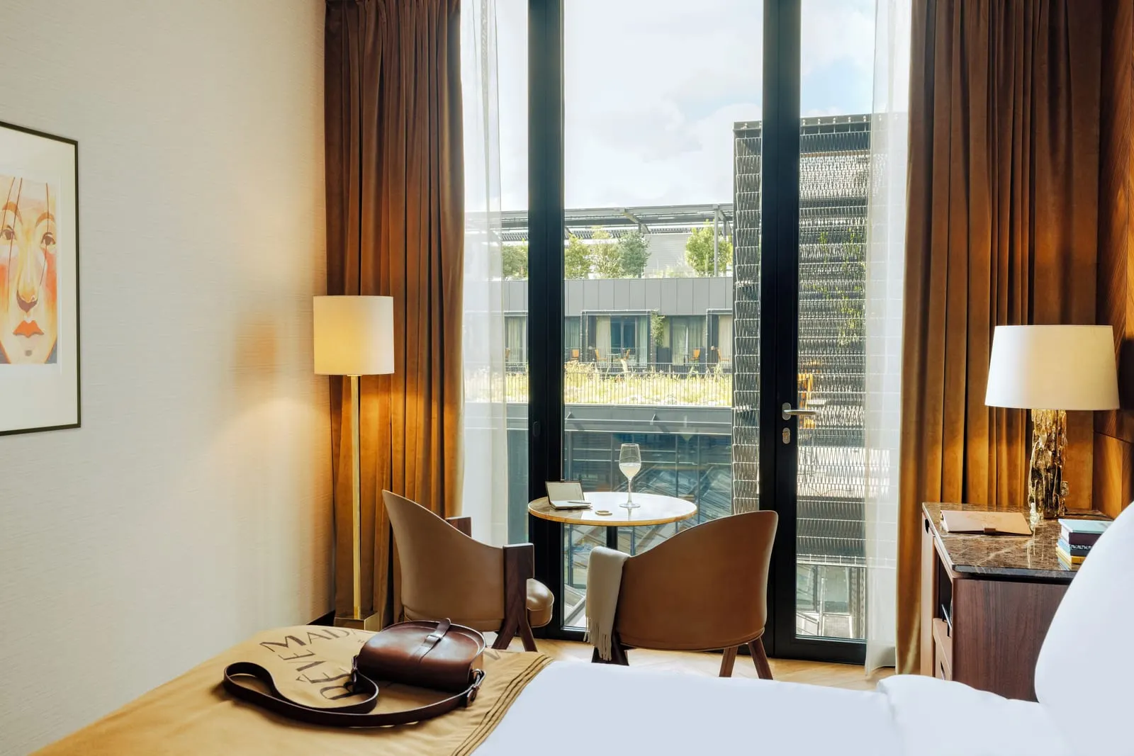 Superior at Hôtel Madame Rêve: two leather tub chairs and a small round table by floor-to-ceiling glass doors hung with velvet curtains, a brass floor lamp and a desk, with the planted courtyard and the garden-side rooms beyond