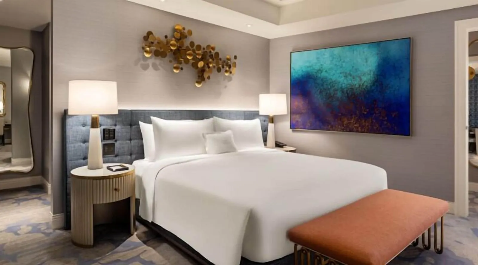 Panorama Suite Mountain View at Fontainebleau Las Vegas: a king bed against a tufted blue headboard under a gold disc sculpture, with a large blue abstract painting and a peach bench at its foot