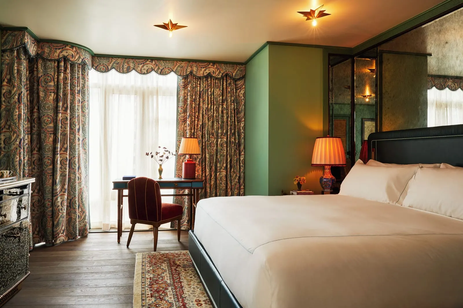 The Corner King at The Fifth Avenue Hotel: a king bed with a dark green headboard, a writing desk and red chair in front of tall windows hung with paisley curtains, and a mirrored wall beside the bed