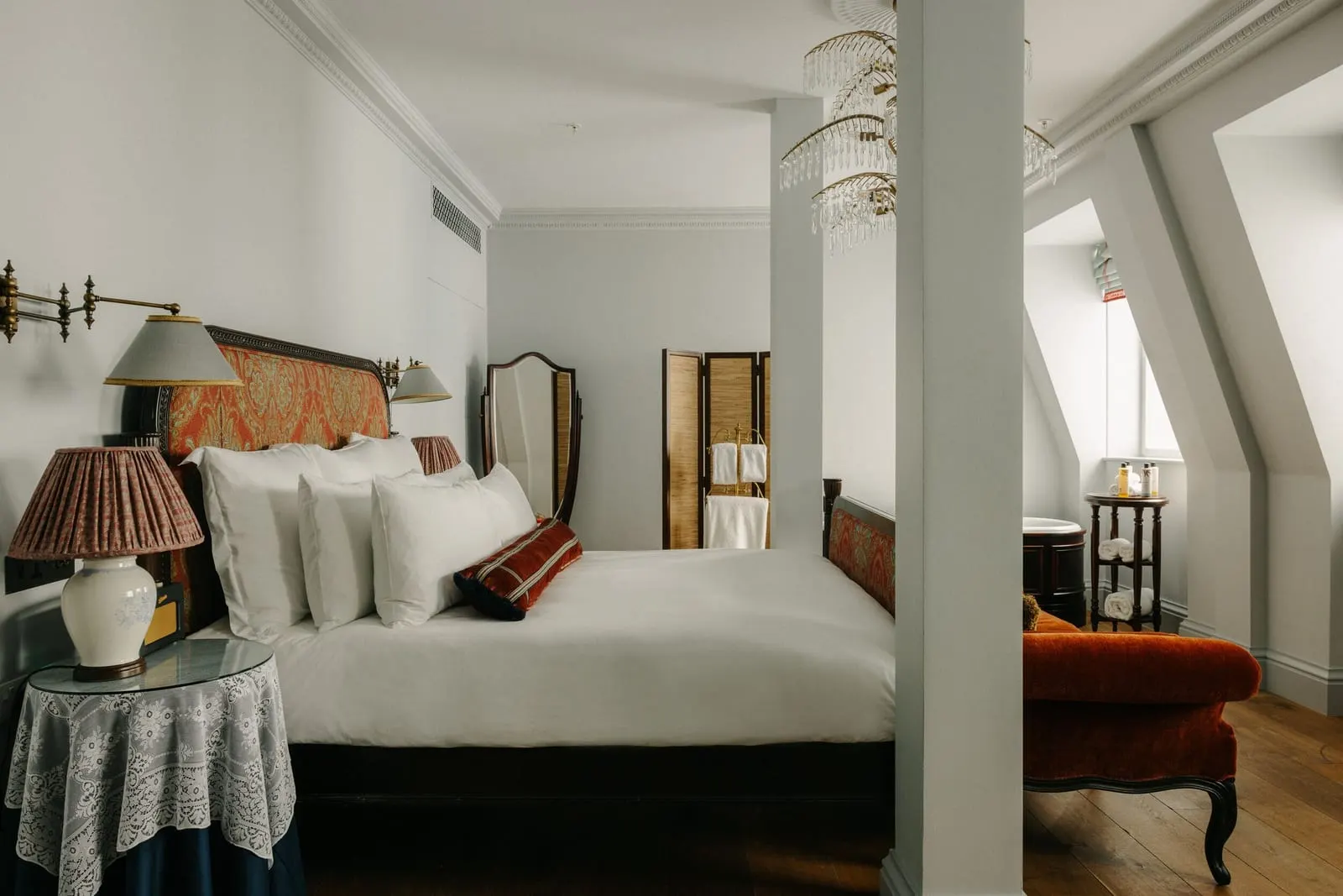 Studio Suite at The Ned: the bed, an orange velvet chaise, a folding screen and a chandelier by angled beams