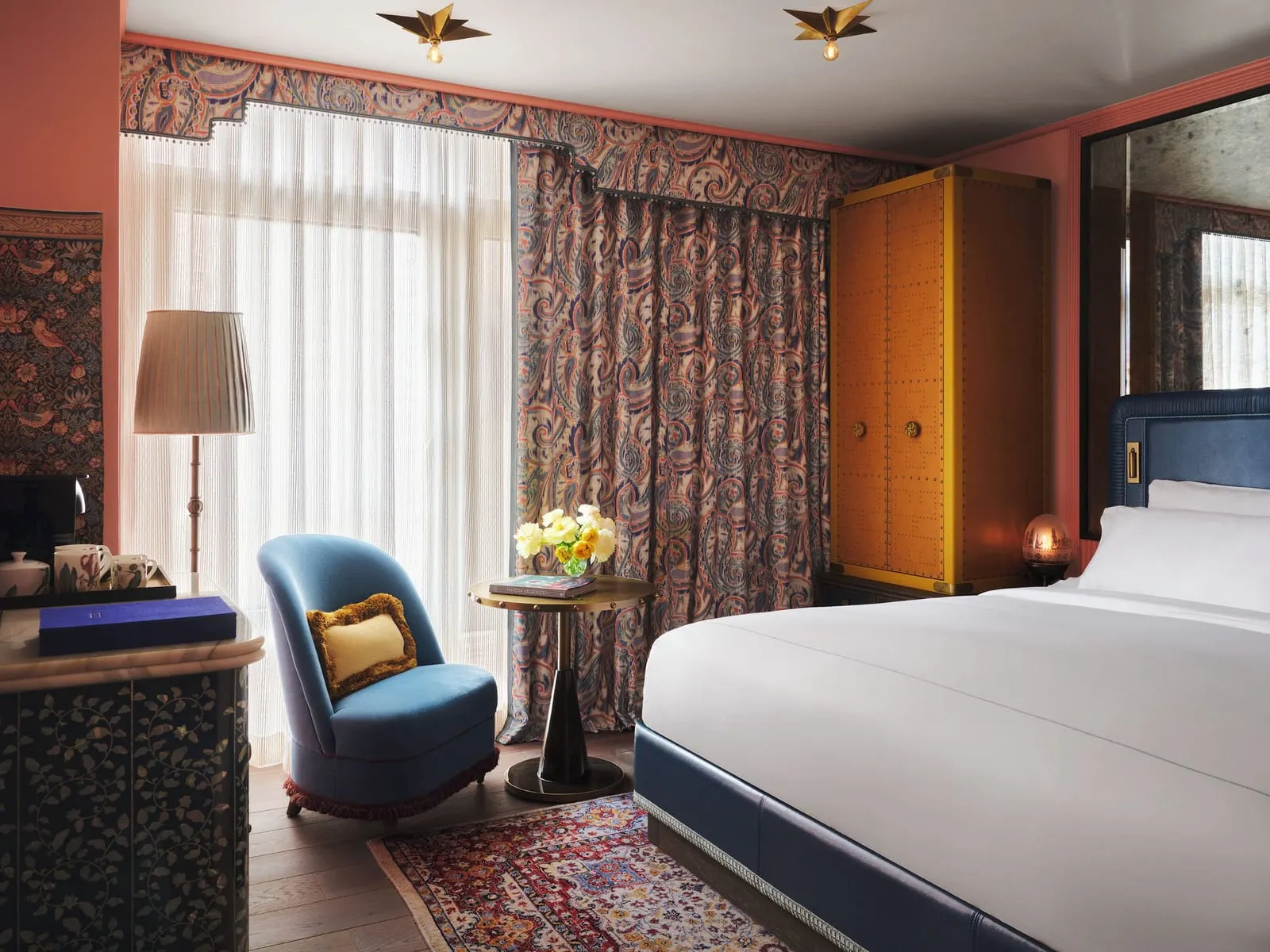 The City King at The Fifth Avenue Hotel: a king bed with white linen and a dark blue headboard, a studded orange wardrobe, a blue armchair and a floor lamp in front of tall sheer curtained windows