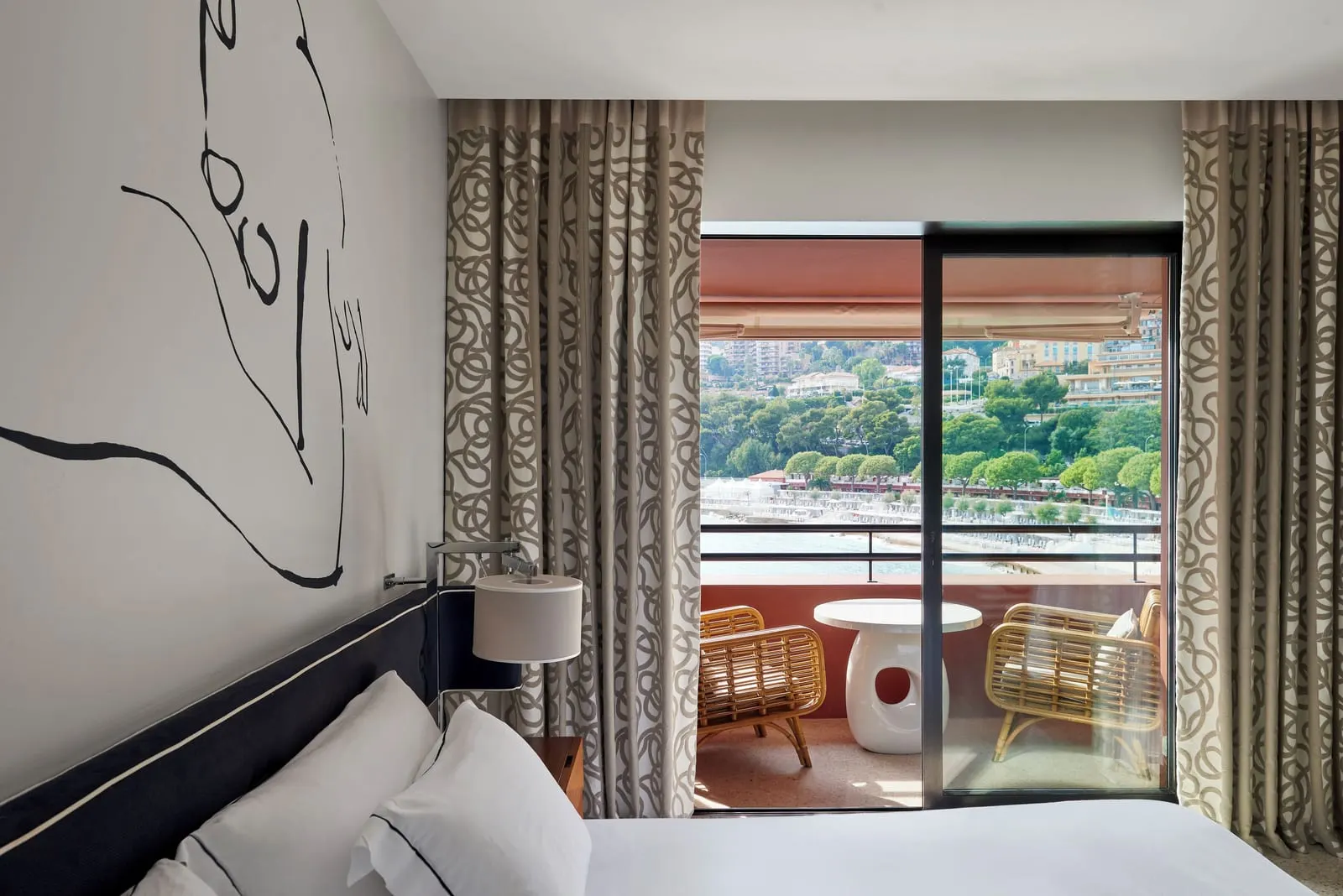 Junior Suite at Monte-Carlo Beach: the bed beside glass doors to a loggia with rattan chairs, the beach and trees beyond