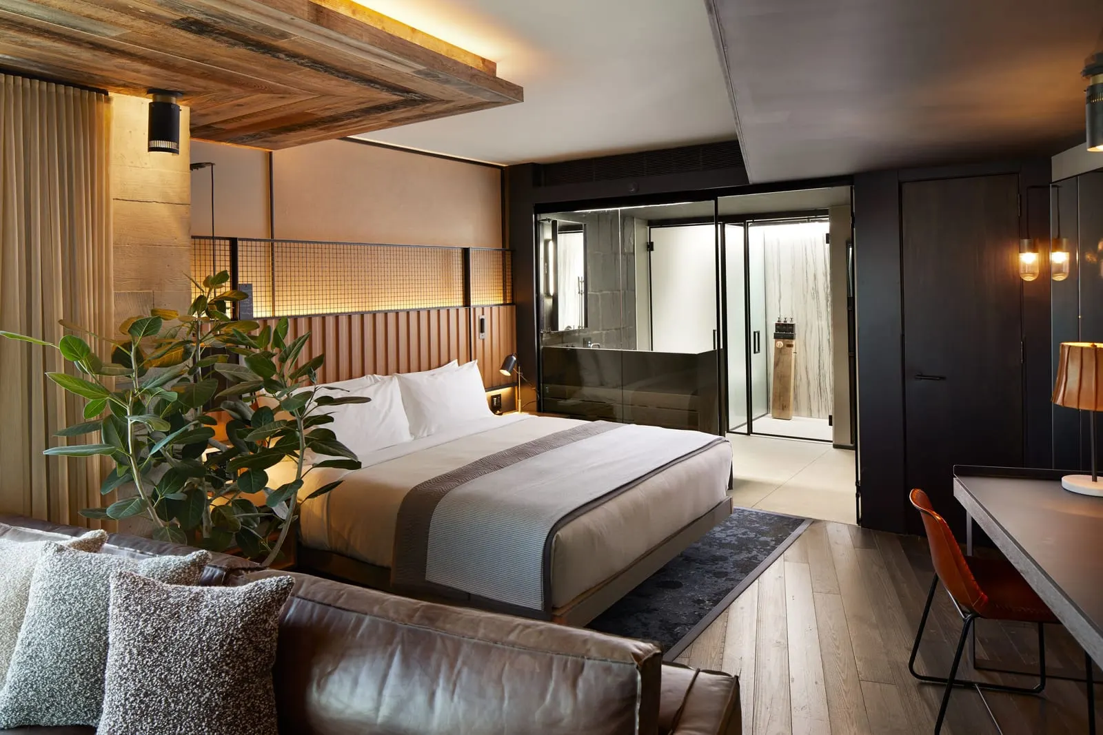Liberty Studio Suite im 1 Hotel Brooklyn Bridge: ein Kingsize-Bett mit einem Ledersofa im Vordergrund, eine große Pflanze, ein Schreibtisch mit einem hellbraunen Lederstuhl und ein Bad mit Glaswänden hinter dem Fußende des Bettes