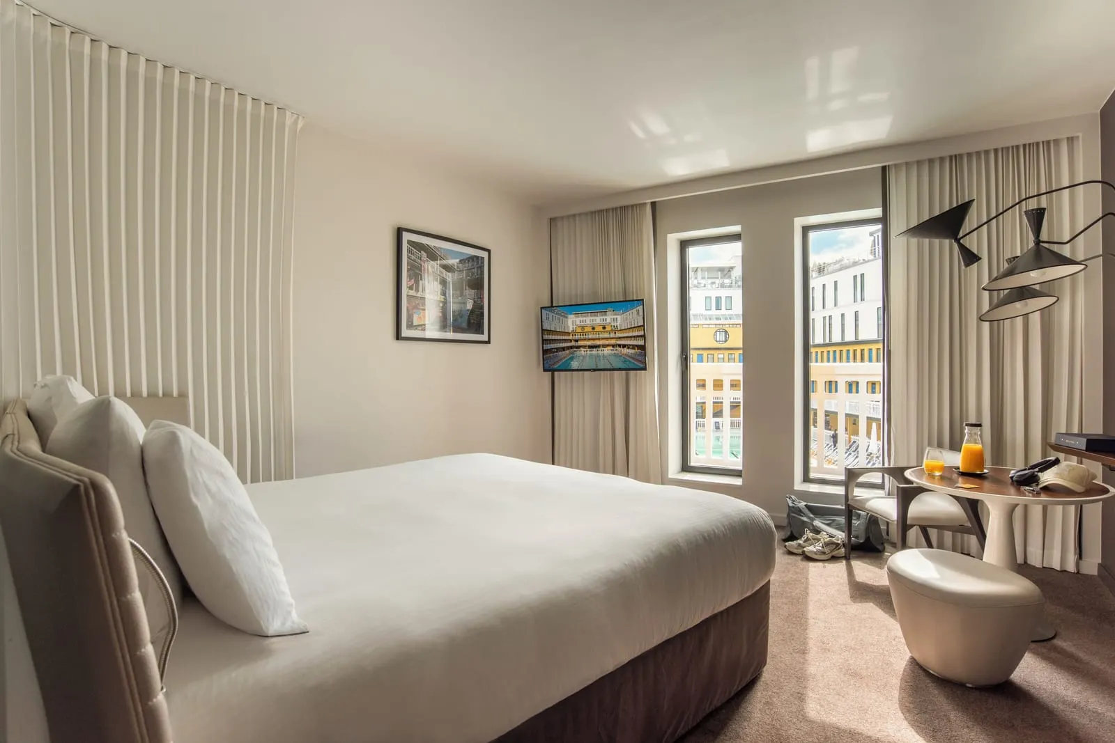 Deluxe Room at Hôtel Molitor Paris: a king-size bed, a small round table with a chair and two tall windows onto the galleries and blue cabin doors around the outdoor pool