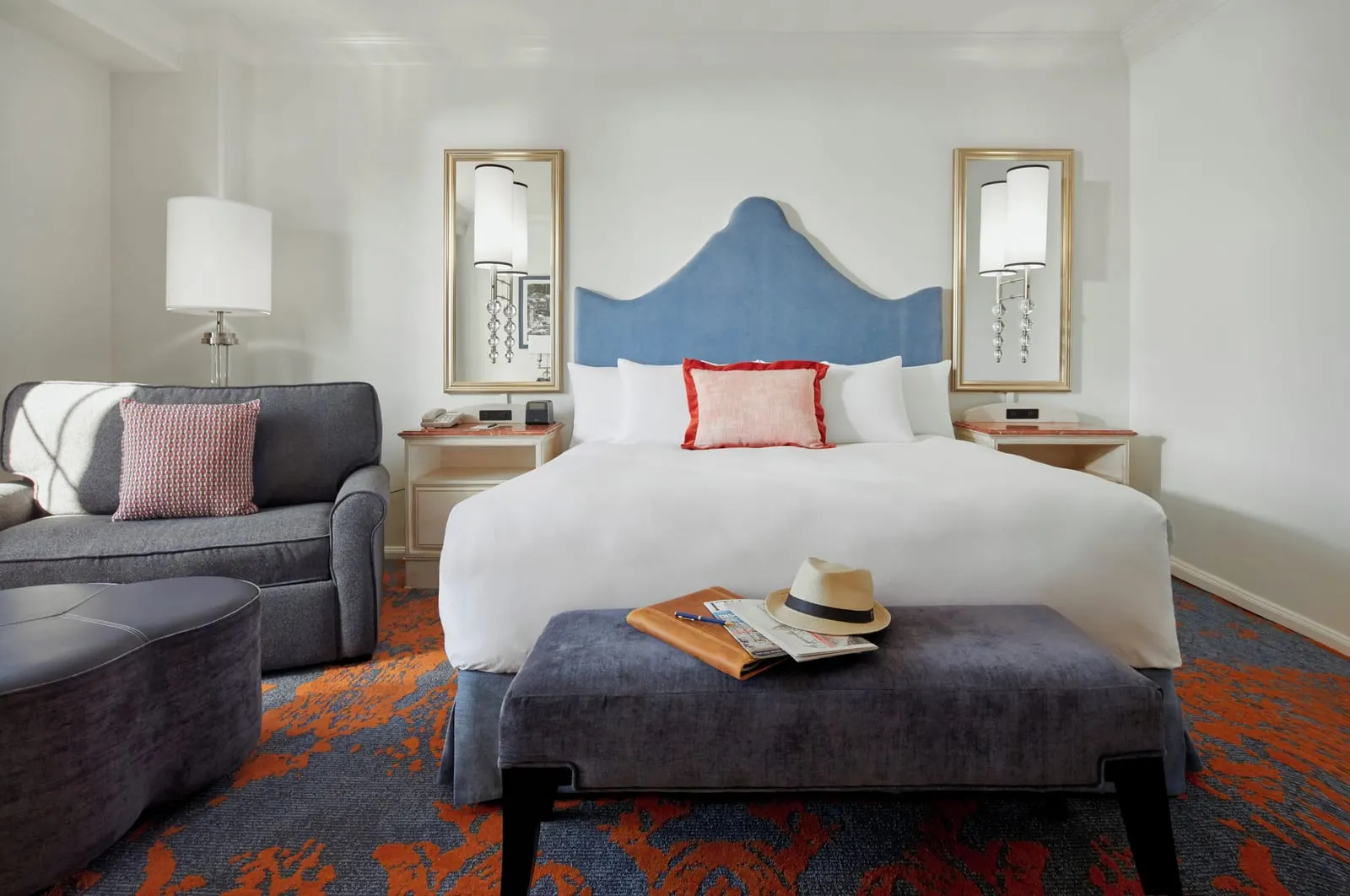 Bay View King Room at Loews Portofino Bay Hotel: the king bed with a blue upholstered headboard, wall lamps mounted on mirrors either side, a bench at its foot and an armchair with an ottoman