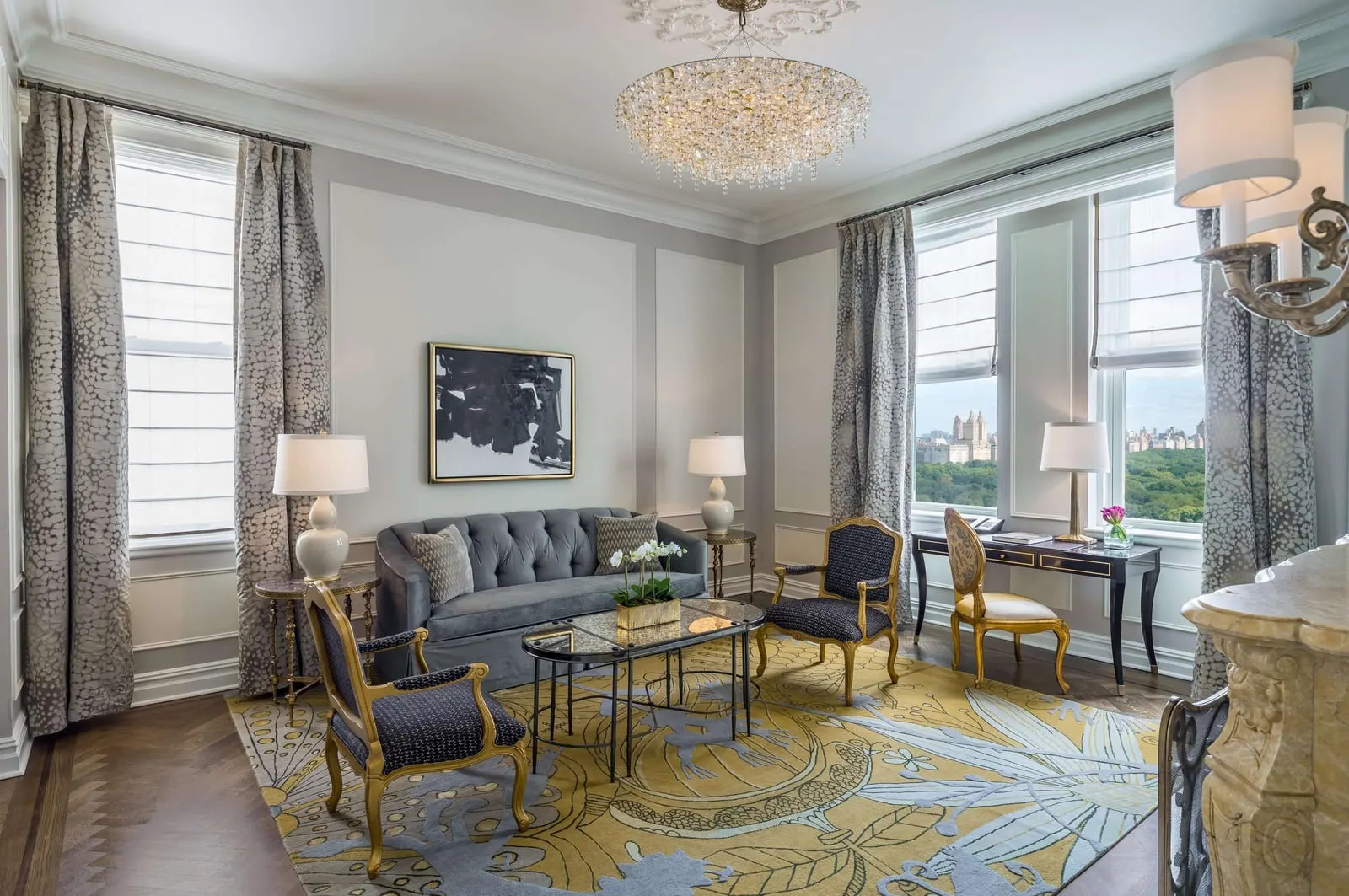 Carnegie Partial Park View One Bedroom Suite King at The Plaza: a sitting room with a grey sofa, gilt armchairs and a glass coffee table, and windows looking over the treetops of Central Park