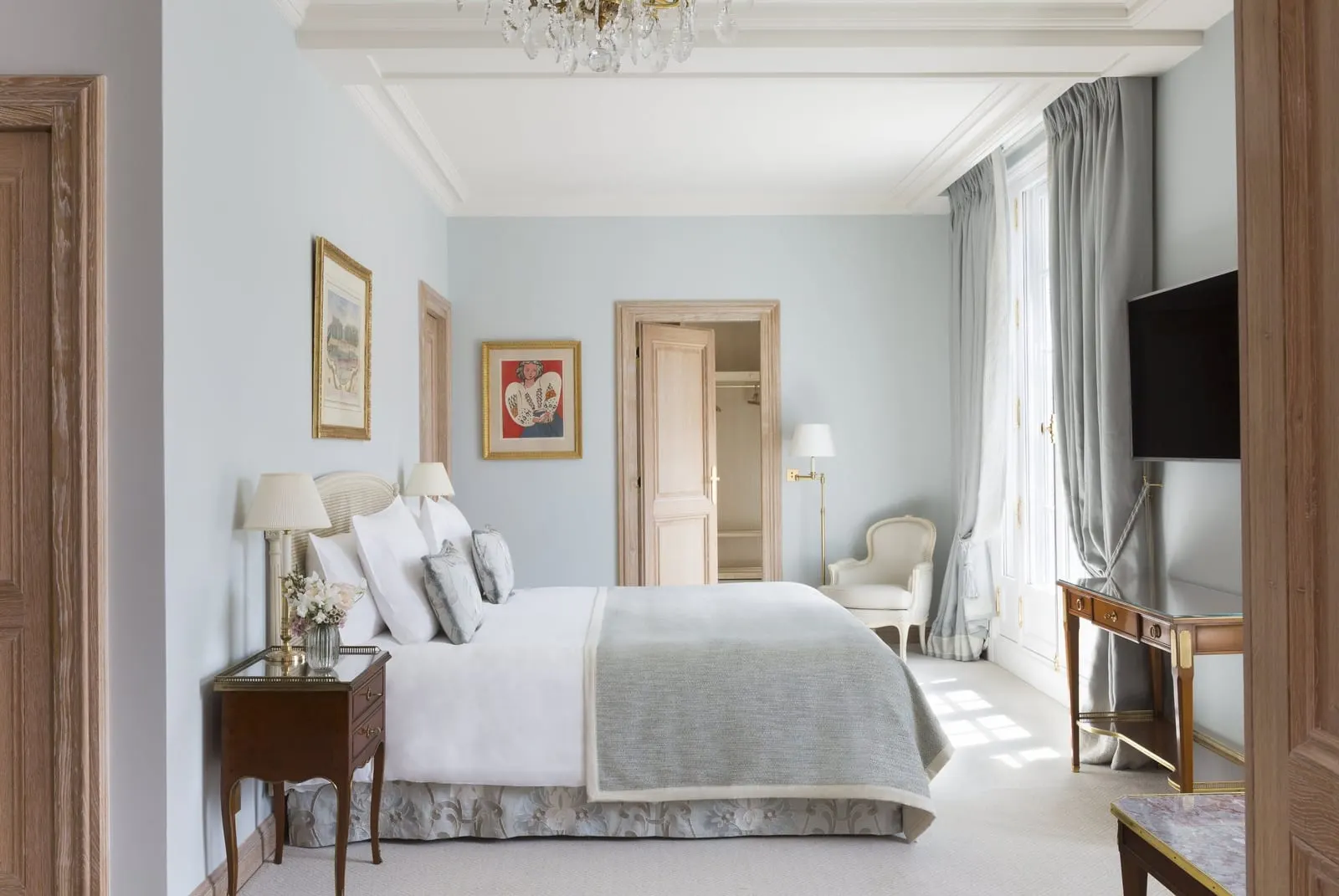 Suite Lumière at Le Bristol Paris: a bed with a grey-blue throw against pale blue walls, framed by light oak door casings, with a writing desk and an armchair by the window