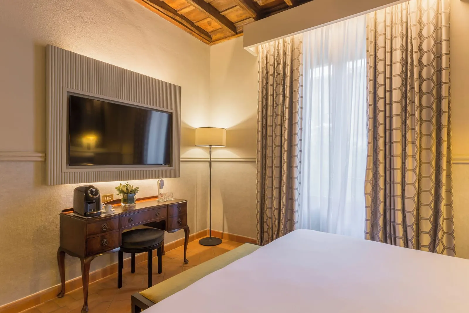 Deluxe Room at Donna Camilla Savelli: the bed under a beamed ceiling, an antique writing desk and a framed TV