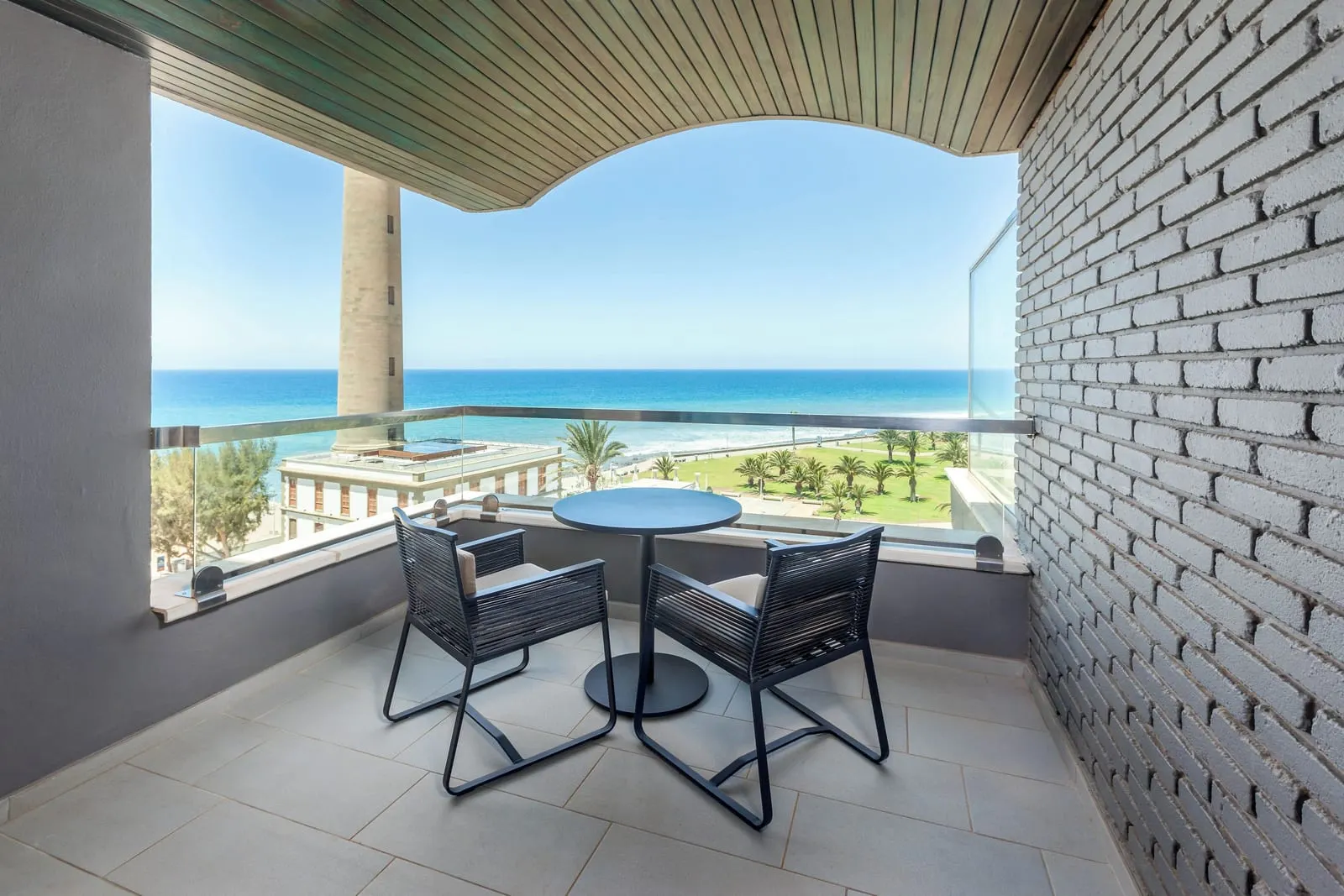 Double Superior at Hotel Faro, a Lopesan Collection Hotel: a balcony with a round table and two chairs beside a grey brick wall, with the lighthouse on the left and the sea beyond