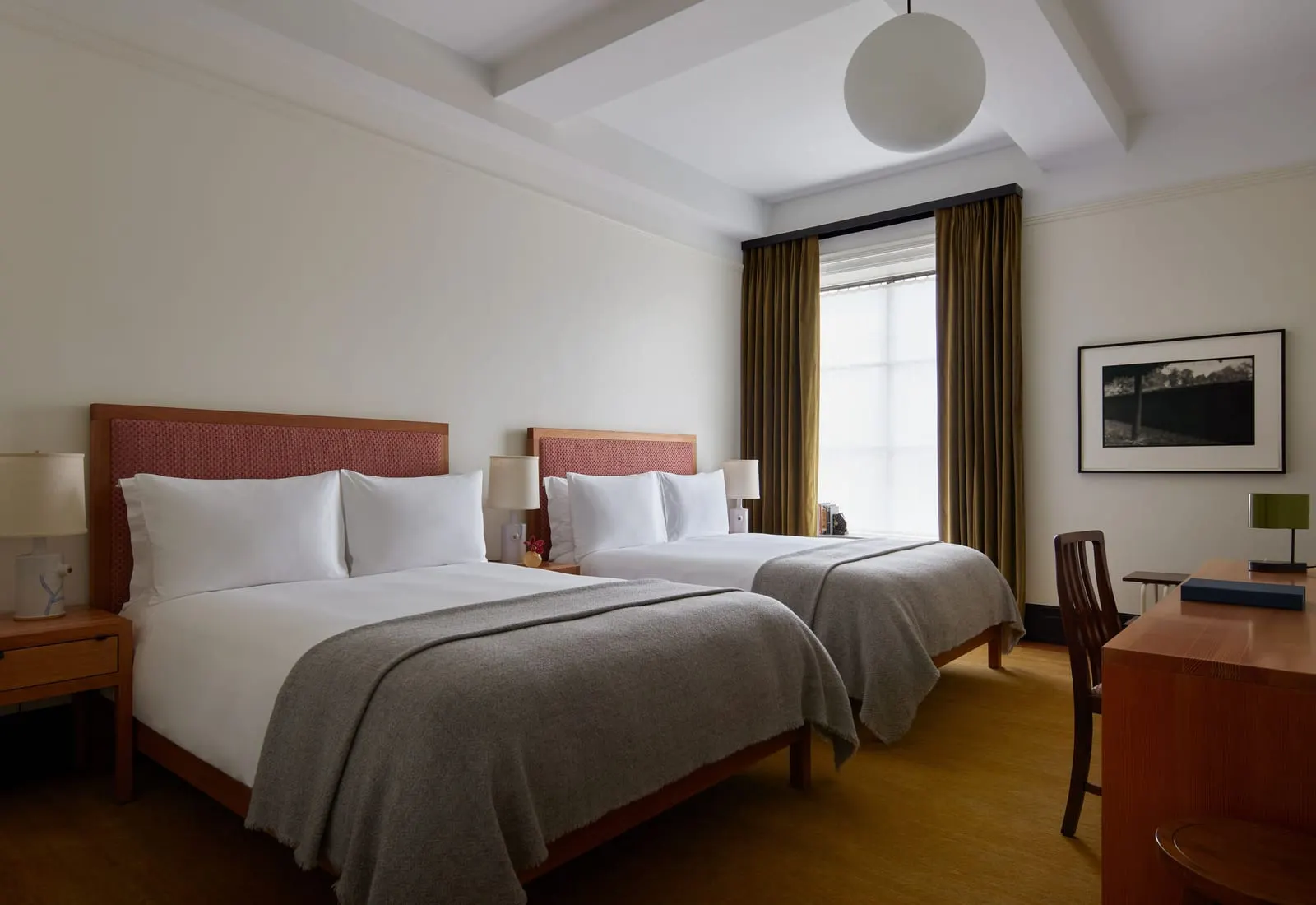 Dual Queen at Nine Orchard: two beds with grey wool throws and woven headboards beside a tall window, with a wooden desk and chair at the right