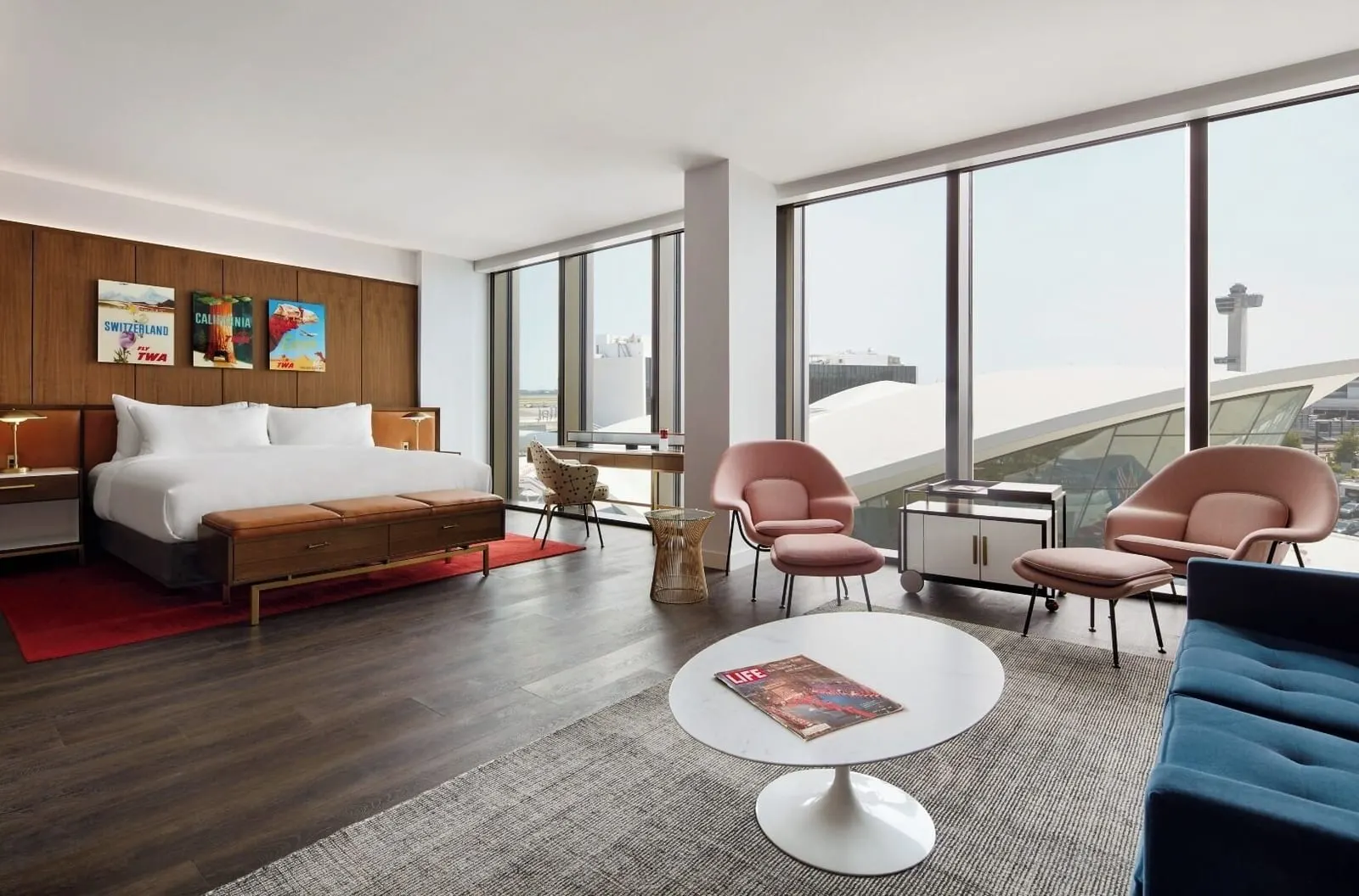 Eero Saarinen Presidential Suite With Historic TWA View at TWA Hotel: a bed with a leather bench on a red rug and a sitting area of pink armchairs, a blue sofa and a round marble table, beside floor-to-ceiling windows facing the TWA Flight Center