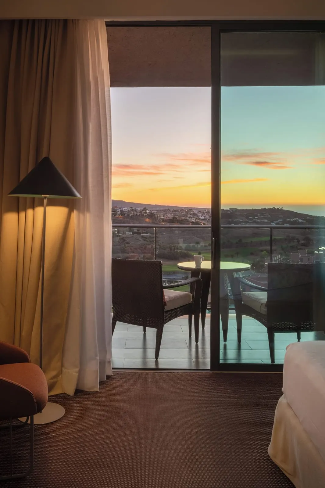 Double Deluxe Superior at Salobre Hotel Resort & Serenity: a carpeted bedroom with a floor lamp, an armchair and the end of the bed, and sliding glass doors to a balcony with two chairs and a table above the golf course at dusk