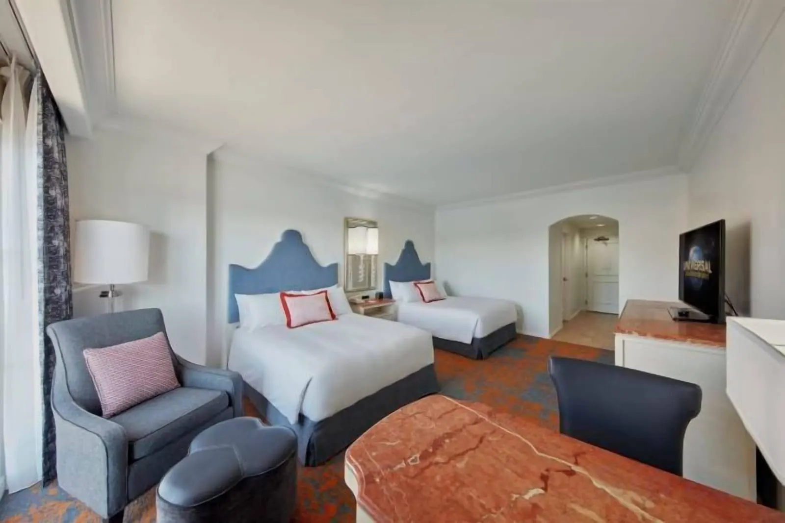 Deluxe 2 Queen Room at Loews Portofino Bay Hotel: two queen beds with blue upholstered headboards, an armchair with an ottoman, a marble-topped desk and a television, with an arched hallway to the door