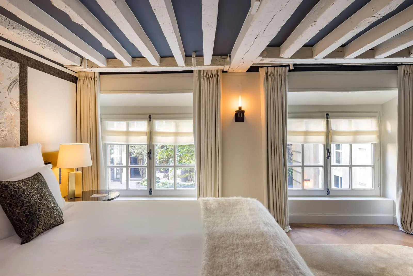 Le Petit Pavillon at Le Pavillon de la Reine: a bedroom under whitewashed beams and a blue ceiling, with a double bed and two deep-set windows