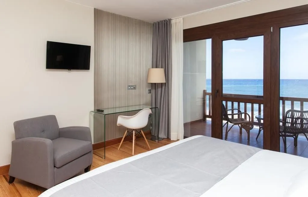 Double Room with Balcony at El Mirador de Fuerteventura: an armchair, glass desk and wall television beside glass doors to a balcony with wicker chairs