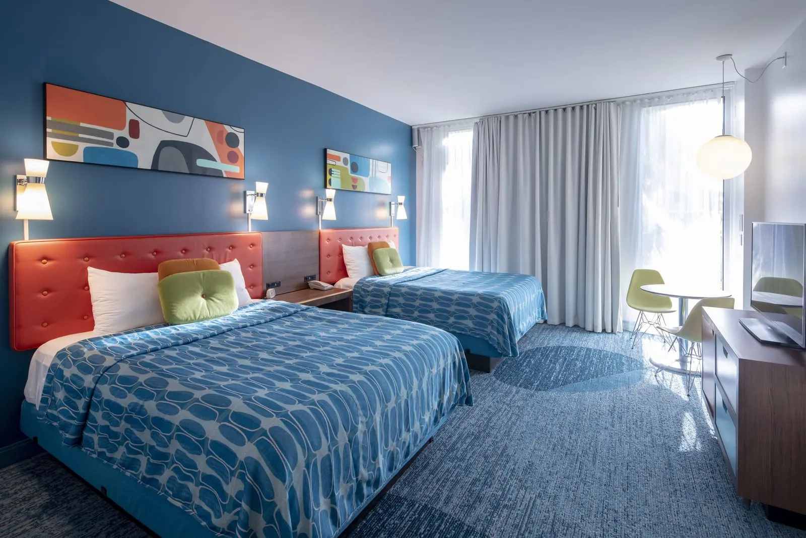 Poolside 2 Queen Beds at Universal Cabana Bay Beach Resort: two queen beds with orange headboards against a dark blue wall, a dresser with a television and a small round table with two chairs by the curtained window