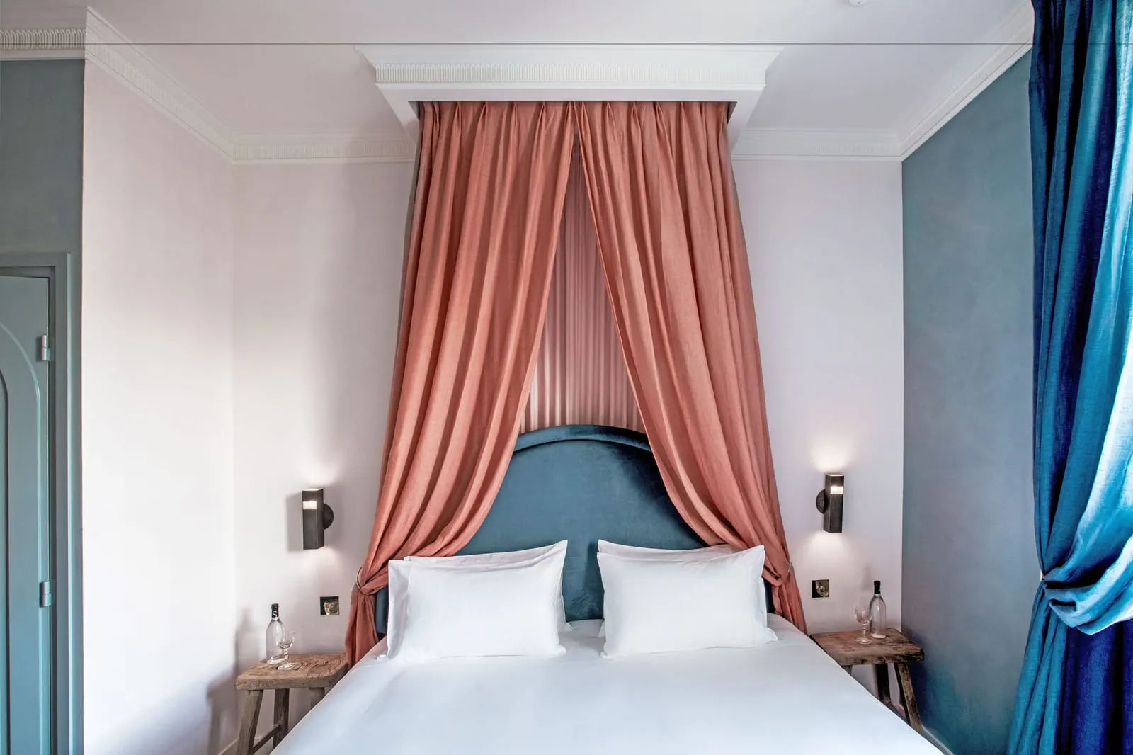 Saint Fiacre Balcony at Grands Boulevards Experimental: a bed under a salmon pink canopy hung from a moulded ceiling box, with a teal velvet headboard, wall lights on each side, a slate-blue wall and a blue curtain drawn back on the right