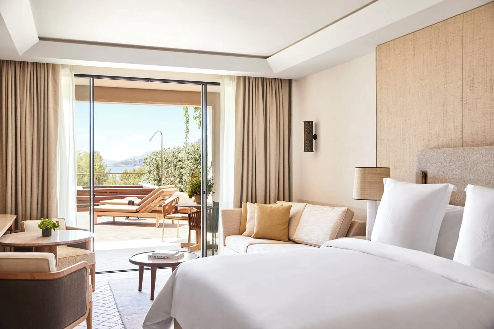 Grand Pine-View Suite with Plunge Pool im Four Seasons Resort Mallorca at Formentor: ein Kingsize-Bett und ein Sofa gegenüber offenen Glastüren zu einer Terrasse mit Sonnenliegen, Bäumen und einem schmalen Meerblick
