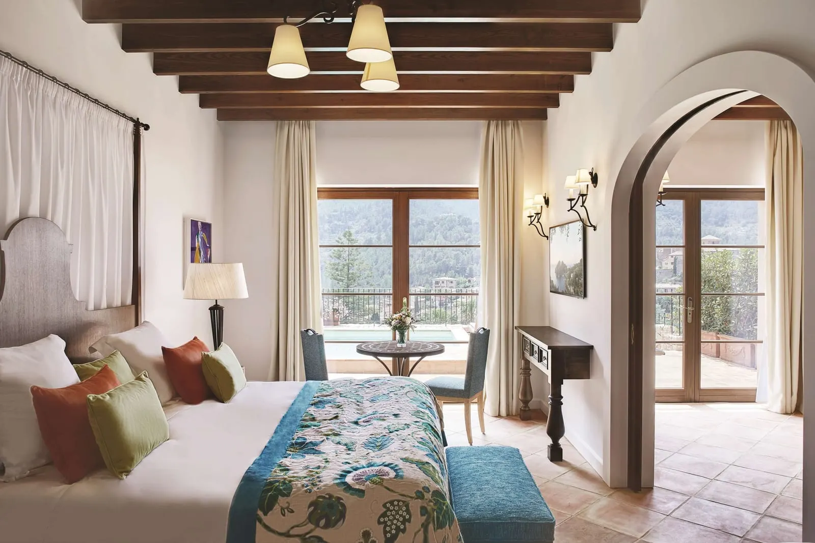 Exclusive Suite at Belmond La Residencia: a bed with a floral runner and a blue bench under wooden beams, glass doors to a terrace with a pool and wooded hills beyond, and an arch to a second room