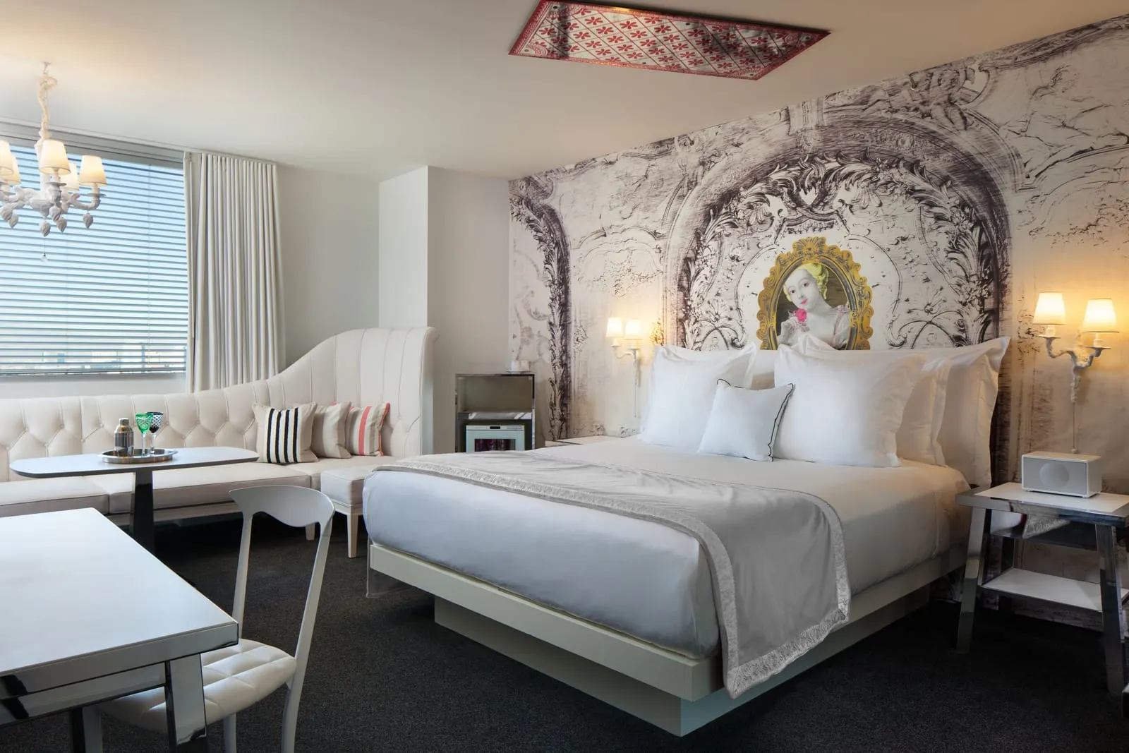 Alexandria Deluxe King at SAHARA Las Vegas: a king bed against a black and white baroque-print wall with a framed portrait, a white tufted banquette and table by the window, and a desk with a white chair