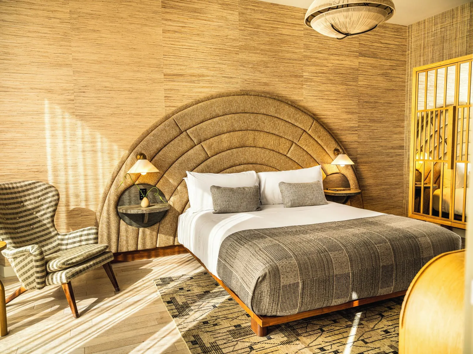 Sunset One Bedroom Suite im Santa Monica Proper Hotel: ein Kingsize-Bett mit einem bogenförmigen Kopfteil vor einer geflochtenen Wandverkleidung, mit einem gestreiften Sessel und einem verspiegelten Raumteiler aus Holz